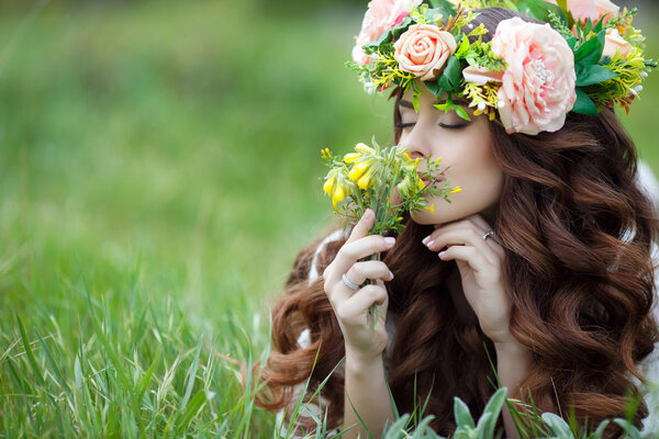 Spring portrait of a beautiful woman in a wreath of flowers,long curly red hair,gray eyes,light makeup and a beautiful smile,dressed in a white summer dress,lying on the soft green grass outdoors in spring