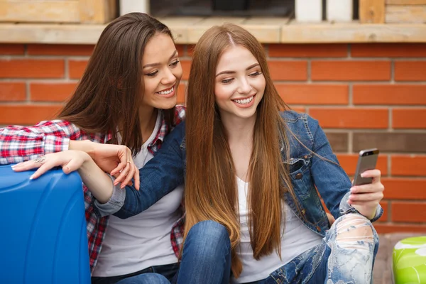 Dos chicas con bolsos leyendo mensajes de texto mientras están sentadas en la estación — Foto de Stock