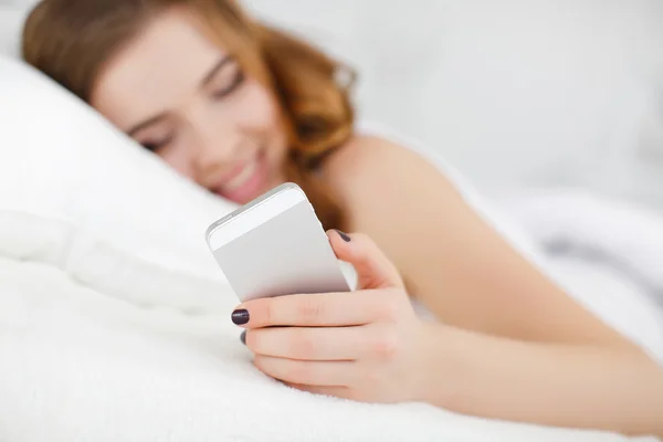 La chica pelirroja leyendo un mensaje en un teléfono inteligente mientras está acostada en la cama — Foto de Stock