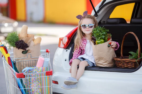 Маленькая девочка-покупатель товаров, сидящая в открытом багажнике автомобиля — стоковое фото