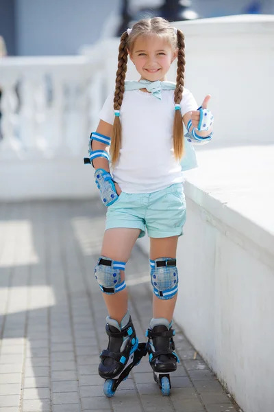 Cute little red-haired girl with hair in two ponytails,grey eyes,dressed in  a multicolored striped dress with short sleeves,learning to roller skate in  the city in the fresh air in the summer Stock