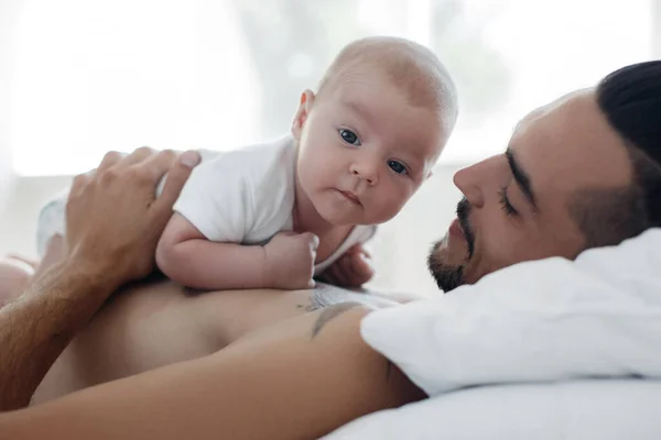 Мужчина с младенцем. счастливая семья дома. счастливый отец с симпатичным двухмесячным мальчиком, лежащим дома на светлой кровати — стоковое фото