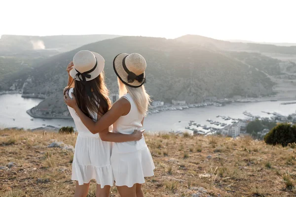 Dois amigos vista traseira ao ar livre, meninas em vestido e chapéu em frente ao mar — Fotografia de Stock
