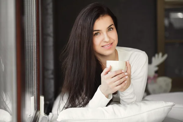 Retrato de una joven acostada en la cama junto a la ventana con una taza de café . — Foto de Stock
