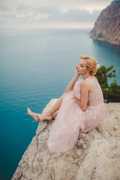 Noiva em vestido de noiva fica em um penhasco com uma bela vista para o mar a partir do topo — Fotografia de Stock