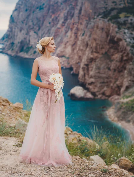 Noiva em vestido de noiva fica em um penhasco com uma bela vista para o mar a partir do topo — Fotografia de Stock