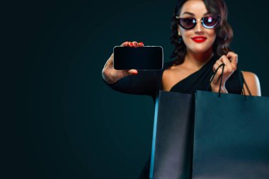 Kara Cuma ve Siber Pazartesi satış konsepti. Yüzünde gülümseme, elinde torbayla ve koyu arka planda izole edilmiş tableti işaret eden alışveriş yapan kadın.