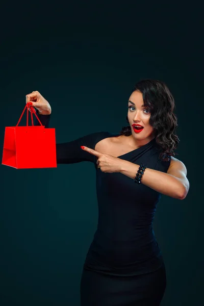 Concept de vente vendredi noir avec belle fille aux cheveux bouclés pour le magasin. Joyeux jeune femme debout isolé sur fond sombre pointant vers la gauche. — Photo