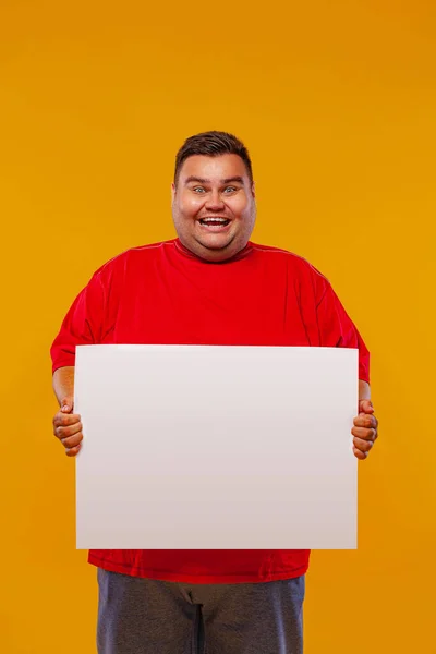 Hombre gordo con camiseta roja mostrando un anuncio de pancarta blanca aislado sobre un fondo amarillo. — Foto de Stock