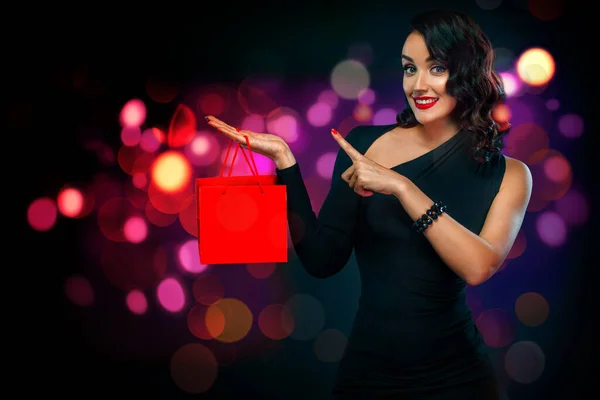 Zwarte vrijdag verkoop concept voor winkel. Shopping girl holding red bag geïsoleerd op donkere achtergrond bij het winkelen op blackfriday. Vrouw wijst naar links kijkend op kopieerruimte. — Stockfoto