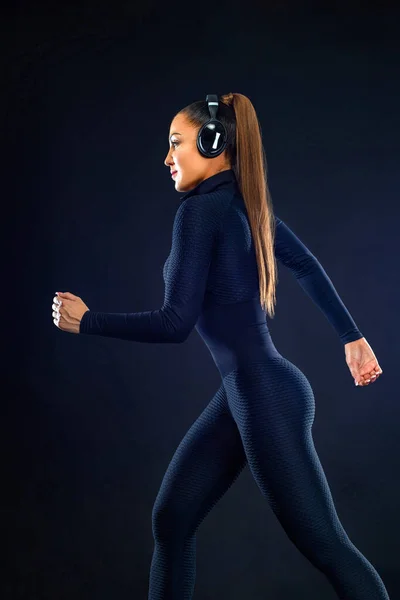 Forte velocista atletica donna, corre su sfondo nero indossando l'abbigliamento sportivo e le cuffie. Fitness e motivazione sportiva. Concetto corridore. — Foto Stock