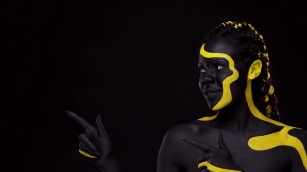 Cara de arte. Muchacha mostrando en el espacio de copia con los dedos. Mujer con pintura corporal negra y amarilla. Chica africana joven con colorido bodypaint. Un increíble modelo afro-americano con maquillaje. — Vídeo de stock