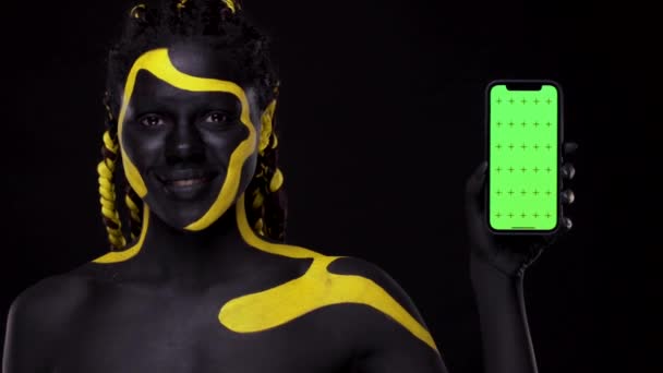 ศิลปะใบหน้า ผู้หญิงที่มีสีผิวดําและสีเหลือง ผู้หญิงชี้นิ้วไปที่หน้าจอโทรศัพท์มือถือด้วยกุญแจโครม่า หญิงสาวชาวแอฟริกันที่มีสีสันทาสีร่างกาย นางแบบแอฟโรอเมริกันที่น่าทึ่งพร้อมเครื่องสําอาง . — วีดีโอสต็อก