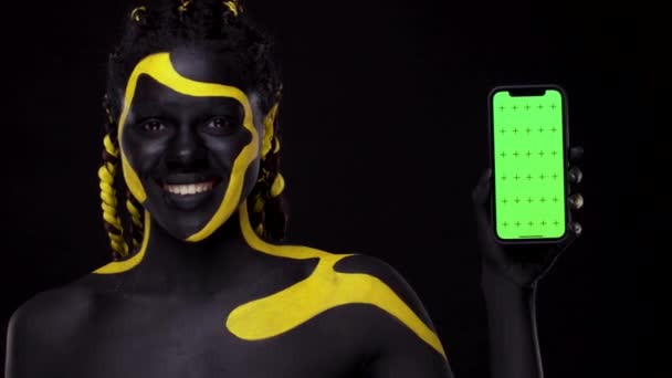 Twarzą w twarz. Kobieta z czarno-żółtą farbą do ciała. Dziewczyna wskazując palcem na ekranie telefonu komórkowego z kluczem chroma. Młoda afrykańska kobieta z kolorowymi farbami do ciała. Niesamowity afro amerykański model z makijażem. — Wideo stockowe