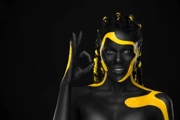 Arcművészet. Egy lány, aki jól mutat ujjal. Nő, fekete és sárga testfestékkel. Fiatal afrikai lány színes testfestékkel. Egy csodálatos afro-amerikai modell sminkkel. — Stock Fotó