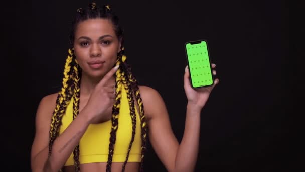 Portret van een lachend Afrikaans meisje met de vinger naar een mobiel telefoonscherm met chroma sleutel. vrouw geïsoleerd over zwarte achtergrond — Stockvideo