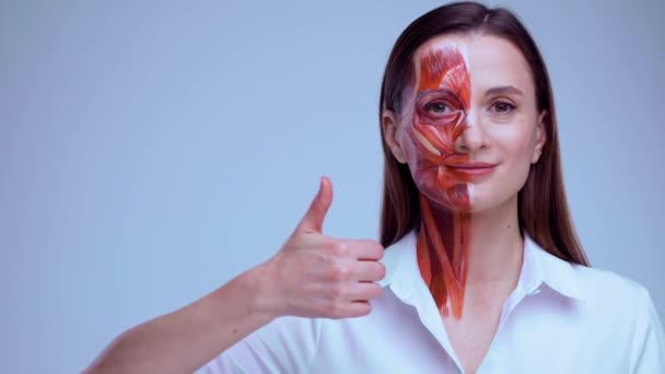 Glückliche junge Frau mit halbem Gesicht und Muskelstruktur unter der Haut mit erhobenen Daumen. Modell für die medizinische Ausbildung auf hellem Hintergrund. Nahaufnahme Video von Gesicht menschliche Anantomie. — Stockvideo