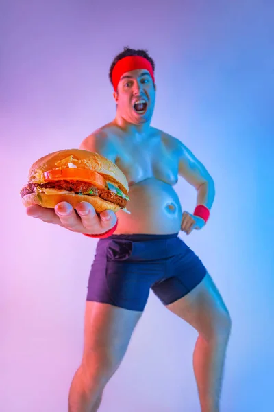 Dicke Mann mit verschenkt Hamburger, um Gewicht zu verlieren und ein schlanker Sportler zu werden. Nicht sportlicher Mann isst Hamburger isoliert auf rosa Hintergrund. — Stockfoto