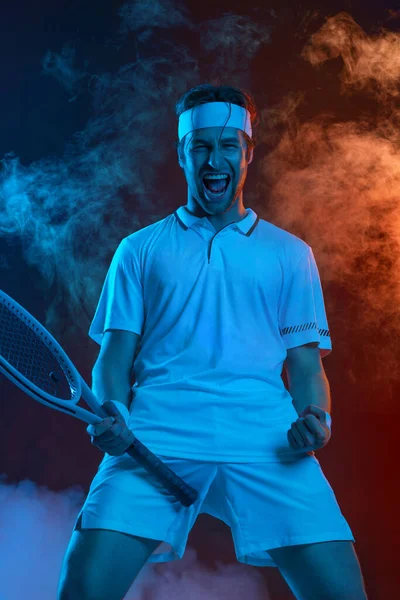 Gelukkig tennisser viert overwinning. Tennis atleet met racket in wit t-shirt. Man spelen op donkere achtergrond met kleur rook. — Stockfoto