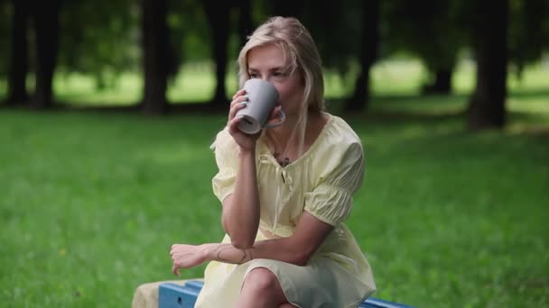 公園のベンチに座っている間 明るい夏の日に美しい女の子はお茶やコーヒーを飲みます スタイリッシュなヒップスターの女の子は屋外のベンチに座っています 彼女は酒を楽しんでいる — ストック動画