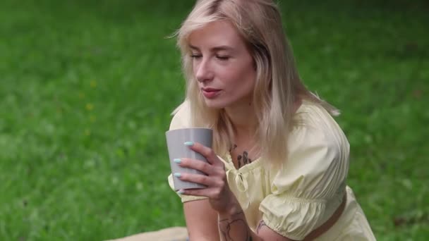 一个美丽的女孩在阳光灿烂的夏天坐在公园的长椅上喝茶或喝咖啡 时髦的姑娘坐在外面的长椅上 她正享受着喝一杯 — 图库视频影像