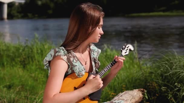 Mladá krásná dívka hraje na hudební nástroj na pozadí řeky. — Stock video