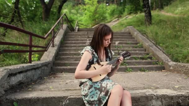 Young beautiful girl playing a musical instrument mandolin while sitting on the steps of the stairs — Stock Video