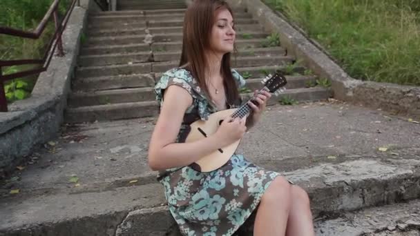 Mladá krásná dívka hraje na hudební nástroj mandolínu, zatímco sedí na schodech — Stock video