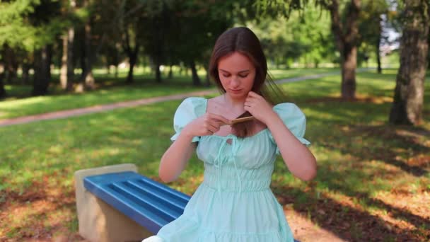 Menina bonita penteando seu cabelo longo enquanto sentada em um parque — Vídeo de Stock