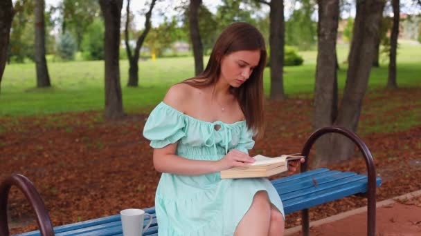 아름다운 소녀는 여름날 벤치에 읽습니다 소녀는 밖에서 마신다 — 비디오