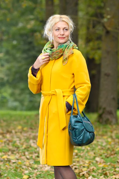 Senior woman in a yellow coat walks in the park — Stock Photo, Image