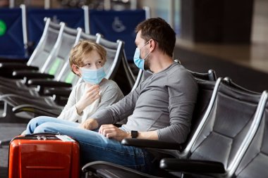 İki kişilik aile, baba ve oğul, yüz maskesi takıyorlar havaalanı terminalinde oturuyorlar, koronavirüs salgını sırasında seyahat ediyorlar.