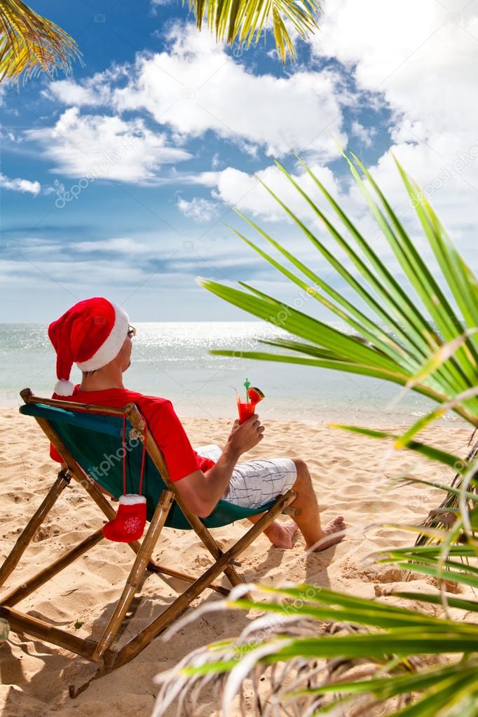 Man in Santa's hat with cocktail sitting on chair on a beach