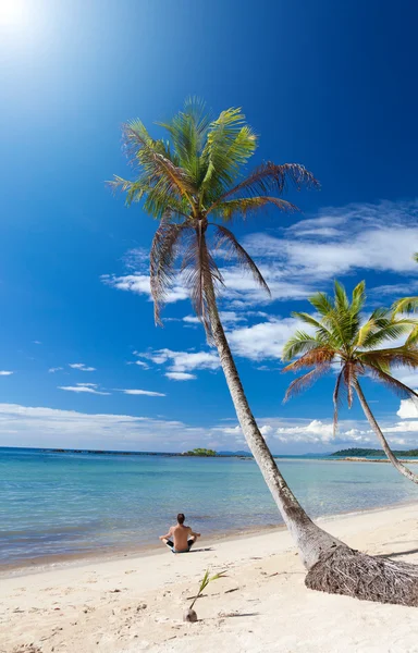 Hombre en una postura de yoga se relaja bajo las palmeras en una playa solitaria — Foto de Stock