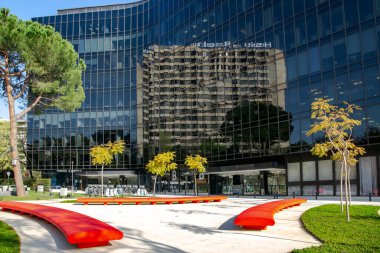 İspanya, BARCELONA, MARCH, 2021: Ön cephesinde koyu cam paneller olan modern ofis binası ve İspanya 'nın Barcelona kentindeki avluda bankları olan güzel bir bahçe