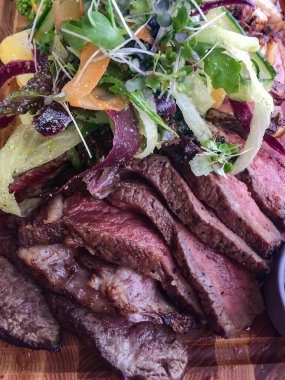 Izgara sulu mermer biftek parçaları, ahşap bir tahtanın üzerinde baharatlar ve taze otlarla yakın plan.