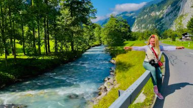 Kıvırcık saçlı bir kadın İsviçre 'nin Bernese Oberland kentindeki bir derenin kenarındaki yol çitlerinde oturuyor. Neşeli seyahat konsepti.