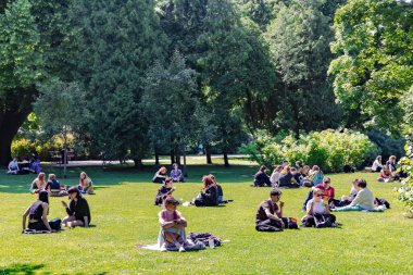 LATVIA, RIGA, JULY, 04, 2025: Arkadaşlarıyla birlikte Riga, Letonya 'daki Central Park' taki bir piknikte çiçek açan çalıların yanındaki yeşil çimlerde oturan aile