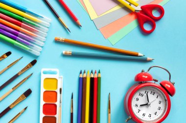 Boyalar, fırçalar, keçeli kalemler, mavi masadaki kalemler. Makas, kağıt ve diğer okul malzemeleri. Kırmızı alarmlı saat üst görünümü.