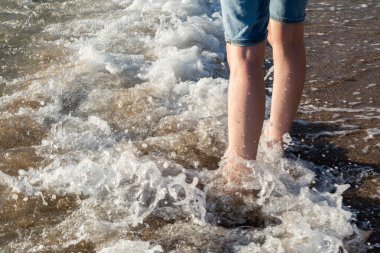 Birisi sahilde duruyor ve yumuşak dalgalar bacaklarına çarpıyor.