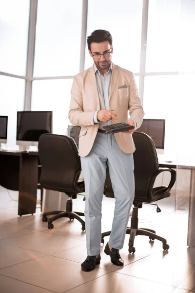 Komoly üzletember számológépet használ, miközben az üres irodában áll — Stock Fotó