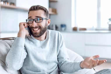 Rahat bir koltukta oturan neşeli bir adam cep telefonuyla konuşuyor.