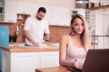 Genç bir kadın mutfakta dizüstü bilgisayarın önünde oturuyor.