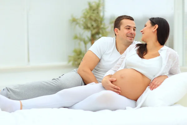 Člověk vypadá láskyplně na svou těhotnou ženu ležící na bílé posteli — Stock fotografie