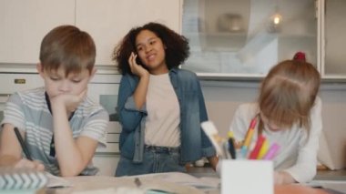 Çocuklar ödevlerini yaparken Afro-Amerikan kadın dadı cep telefonuyla sohbet ediyor.
