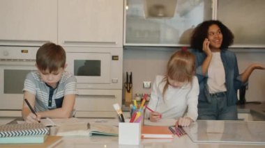 Neşeli bebek bakıcısı cep telefonuyla konuşuyor ve çocuklar ödevlerini yaparken gülüyor.