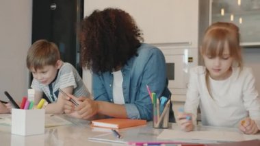 Yardımsever bebek bakıcısı ev ödevi yapan ve evde çizim yapan çocuklarla konuşuyor.