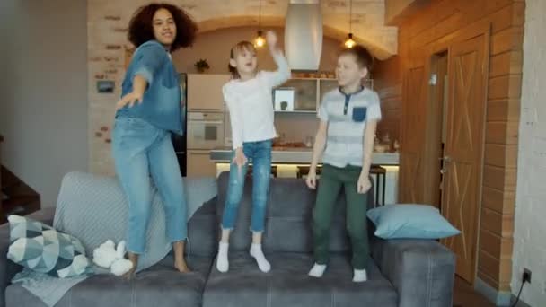 Happy children boy and girl dancing and jumping on sofa with joyful young nanny — Stock Video