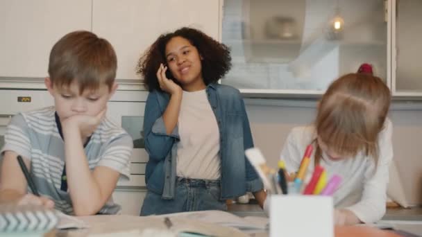 Zorgeloze Afro-Amerikaanse dame nanny chatten op mobiele telefoon terwijl kinderen huiswerk maken — Stockvideo