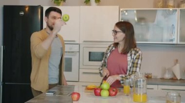 Neşeli adam elma hokkabazlığı yaparken hamile kadın mutfakta meyve kesiyor.